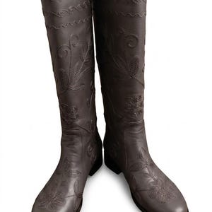 Puede incluir: Un par de botas de cuero marr&oacute;n oscuro hasta la rodilla. Las botas presentan intrincados bordados florales y costuras decorativas. Tienen una punta redondeada y un tac&oacute;n bajo.
