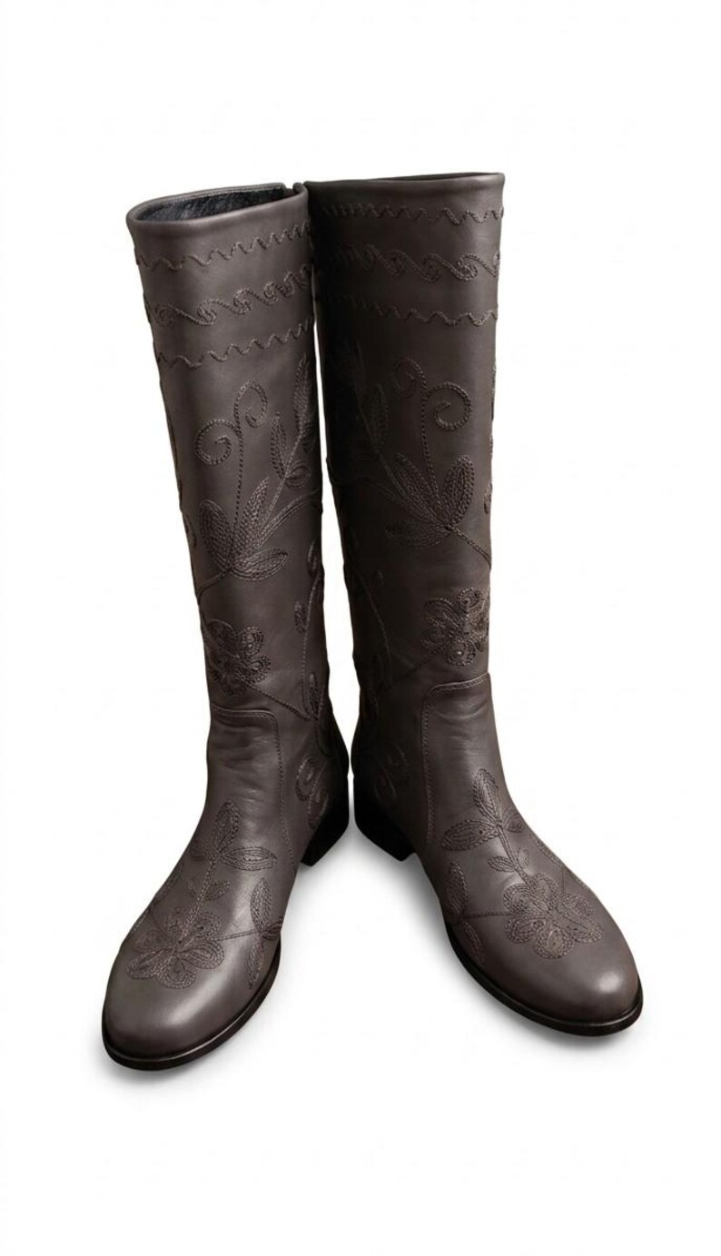 Puede incluir: Un par de botas de cuero marr&oacute;n oscuro hasta la rodilla. Las botas presentan intrincados bordados florales y costuras decorativas. Tienen una punta redondeada y un tac&oacute;n bajo.
