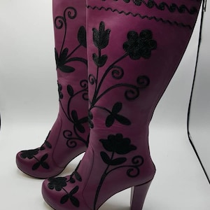 Puede incluir: Un par de botas altas de cuero morado con bordados negros de motivos florales y enredaderas. Las botas tienen punta redondeada, tac&oacute;n de bloque y cierre de cremallera lateral. El bordado se extiende desde la puntera hasta la parte superior.