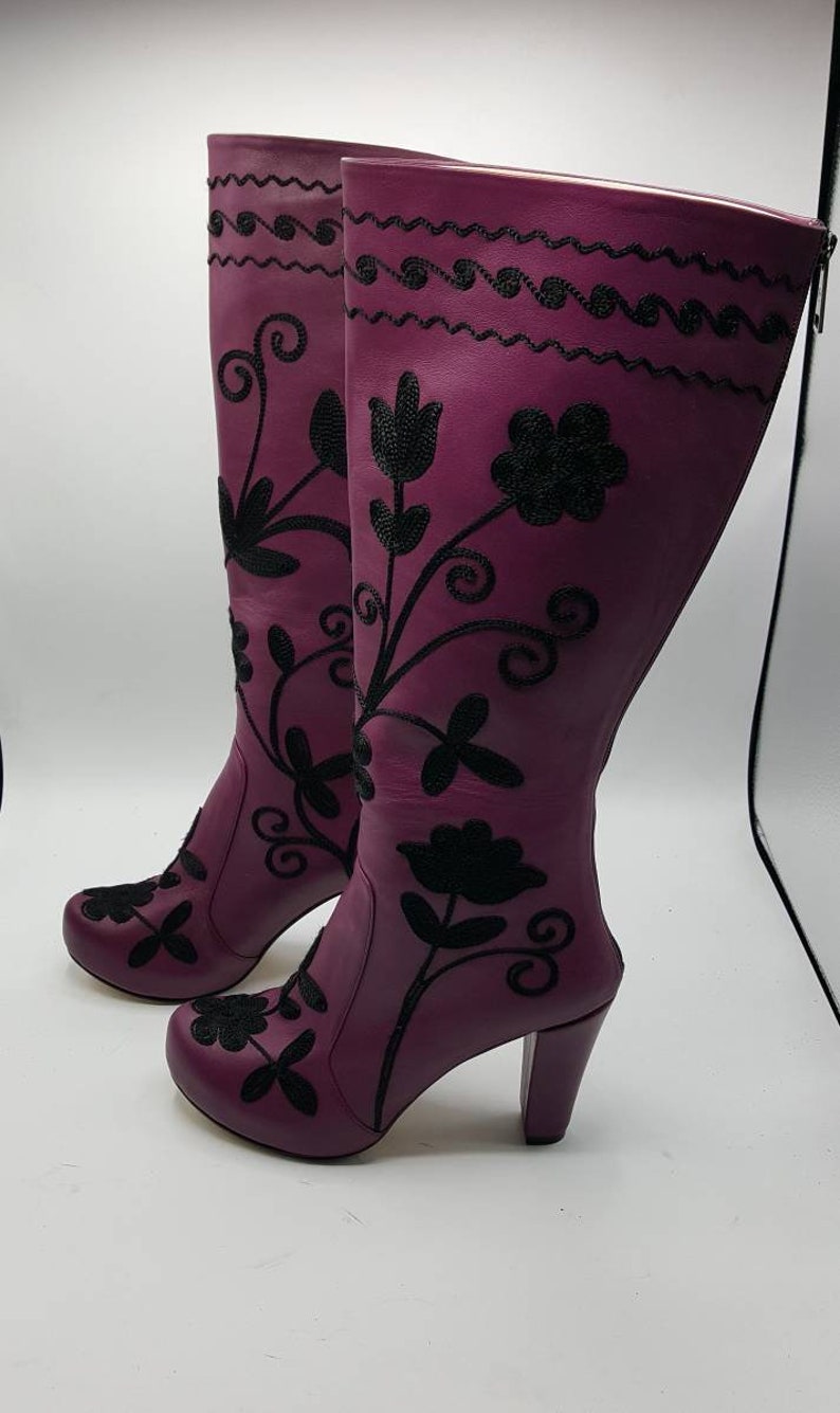 Puede incluir: Un par de botas altas de cuero morado con bordados negros de motivos florales y enredaderas. Las botas tienen punta redondeada, tac&oacute;n de bloque y cierre de cremallera lateral. El bordado se extiende desde la puntera hasta la parte superior.