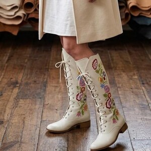 May include: Cream-colored, lace-up knee-high boots with colorful floral embroidery. The boots have a low heel and are worn with a white dress and a cream-colored coat. The boots are the focal point, showcasing a bohemian style.