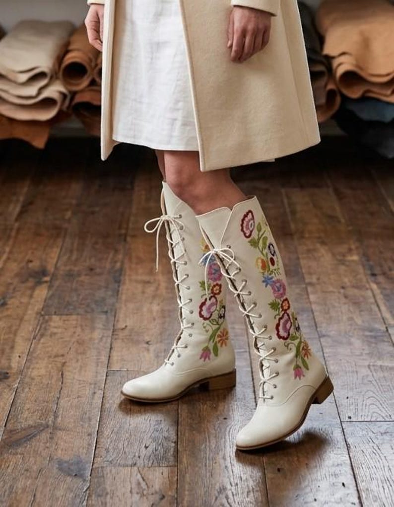 May include: Cream-colored, lace-up knee-high boots with colorful floral embroidery. The boots have a low heel and are worn with a white dress and a cream-colored coat. The boots are the focal point, showcasing a bohemian style.
