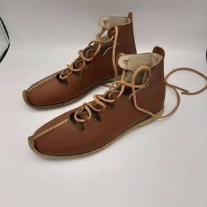 May include: Brown leather lace-up sandals with a cut-out design. The sandals have a flat sole and are tied at the ankle.