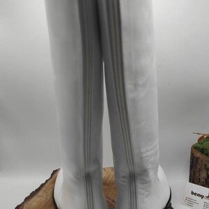 Puede incluir: Un par de botas blancas de cuero de ca&ntilde;a alta con cremallera en la parte posterior. Las botas tienen una suela negra.