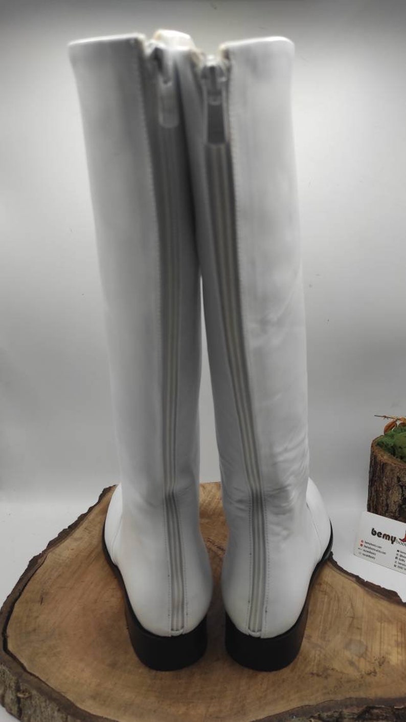 Puede incluir: Un par de botas blancas de cuero de ca&ntilde;a alta con cremallera en la parte posterior. Las botas tienen una suela negra.