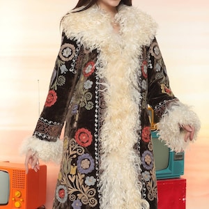 May include: A long, dark brown coat with colorful floral embroidery and cream-colored faux fur trim. The coat features a wide collar, cuffs, and a vertical trim of the same fur. The coat is a statement piece, perfect for a vintage or bohemian style.