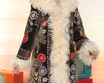 May include: A long, dark brown coat with colorful floral embroidery and cream-colored faux fur trim. The coat features a wide collar, cuffs, and a vertical trim of the same fur. The coat is a statement piece, perfect for a vintage or bohemian style.