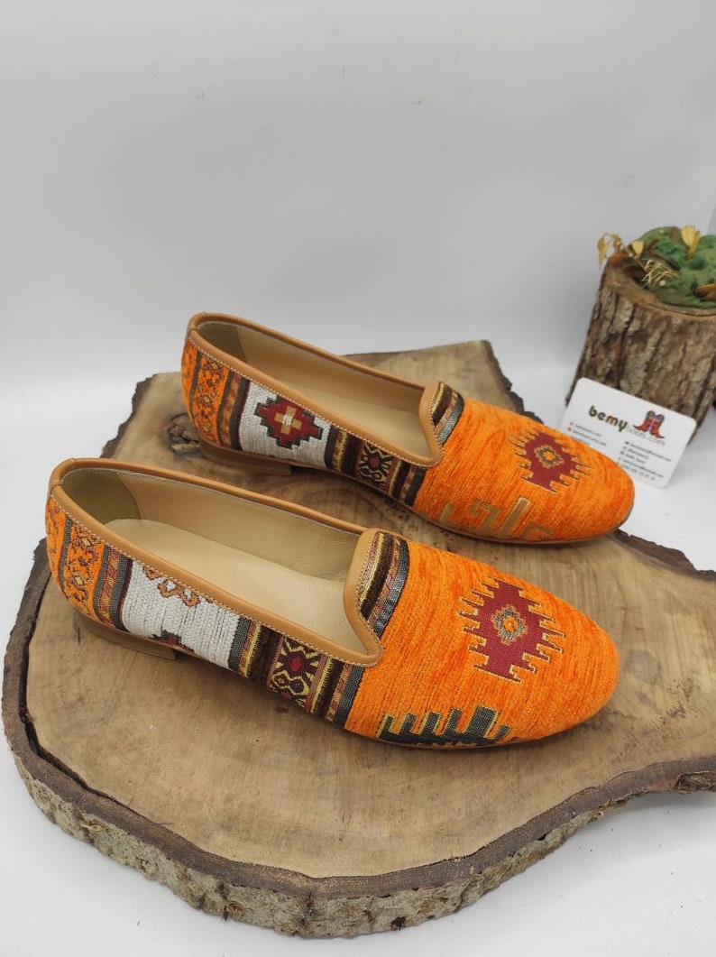 May include: A pair of orange slip-on shoes with a patterned textile design. The shoes have a tan leather trim and a decorative pattern in shades of red, brown, and white. The shoes are displayed on a wooden surface.