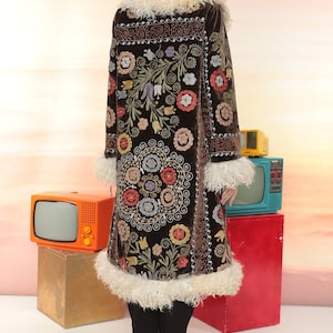 May include: A long, dark brown coat with intricate floral embroidery in various colors. The coat features a cream-colored faux fur collar and trim. The model is wearing black pants and heels.