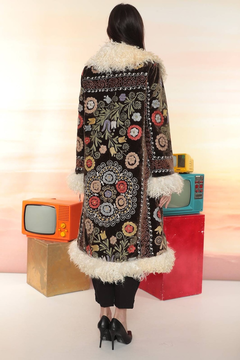 May include: A long, dark brown coat with intricate floral embroidery in various colors. The coat features a cream-colored faux fur collar and trim. The model is wearing black pants and heels.