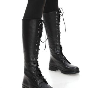 Puede incluir: Botas negras hasta la rodilla con diseño de cordones. Las botas tienen punta redonda y suela gruesa. Están hechas de un material liso similar al cuero y se usan con leggings negros.