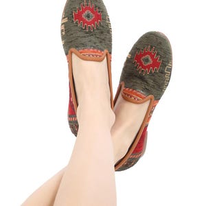 May include: Pair of green and red patterned slip-on shoes. The shoes feature a geometric design in red and brown on the top, with a brown leather trim. The shoes are shown on a white background.