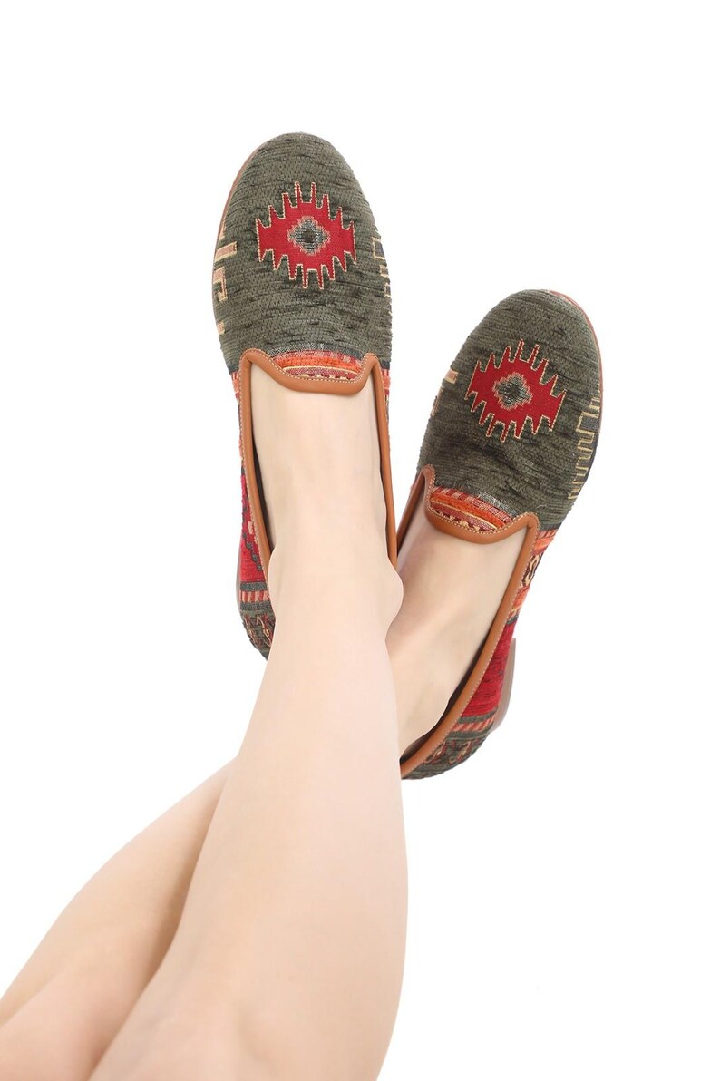 May include: Pair of green and red patterned slip-on shoes. The shoes feature a geometric design in red and brown on the top, with a brown leather trim. The shoes are shown on a white background.