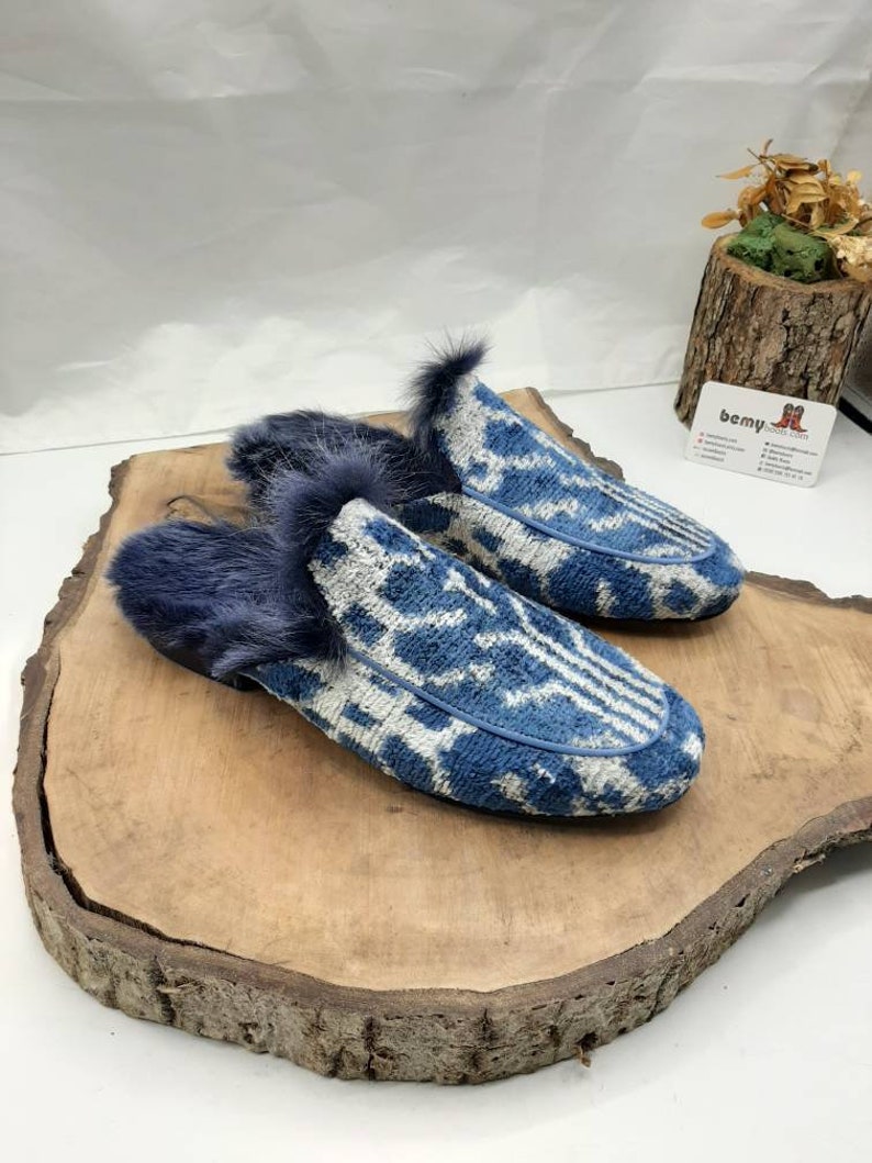May include: A pair of blue and white patterned slippers with a fuzzy gray trim. The slippers are sitting on a wooden surface.
