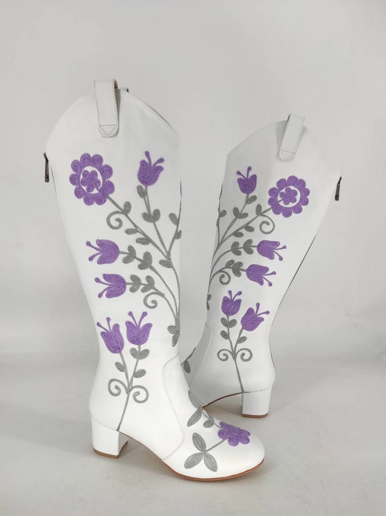May include: White leather knee-high boots with a block heel and purple floral embroidery.