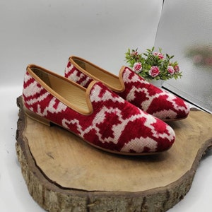 May include: A pair of red and white patterned velvet loafers with a brown leather sole. The loafers are sitting on a wooden surface.
