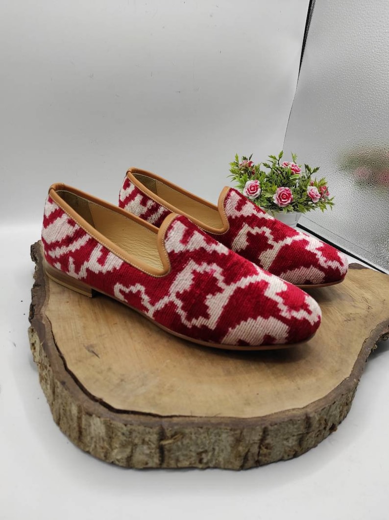 May include: A pair of red and white patterned velvet loafers with a brown leather sole. The loafers are sitting on a wooden surface.