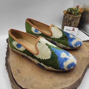May include: A pair of green, blue, and white patterned velvet loafers with a brown leather trim. The loafers are on a wooden surface.