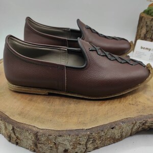 May include: A pair of brown leather slip-on shoes with a braided design on the top. The shoes are on a wooden surface.