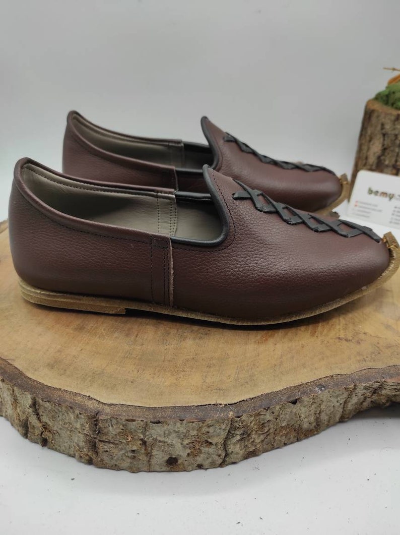 May include: A pair of brown leather slip-on shoes with a braided design on the top. The shoes are on a wooden surface.