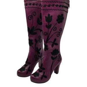 Puede incluir: Un par de botas altas de cuero morado con bordados florales negros y dise&ntilde;os de enredaderas. Las botas tienen punta redondeada, tac&oacute;n de bloque y un cierre de cremallera vertical en la parte posterior de la ca&ntilde;a.