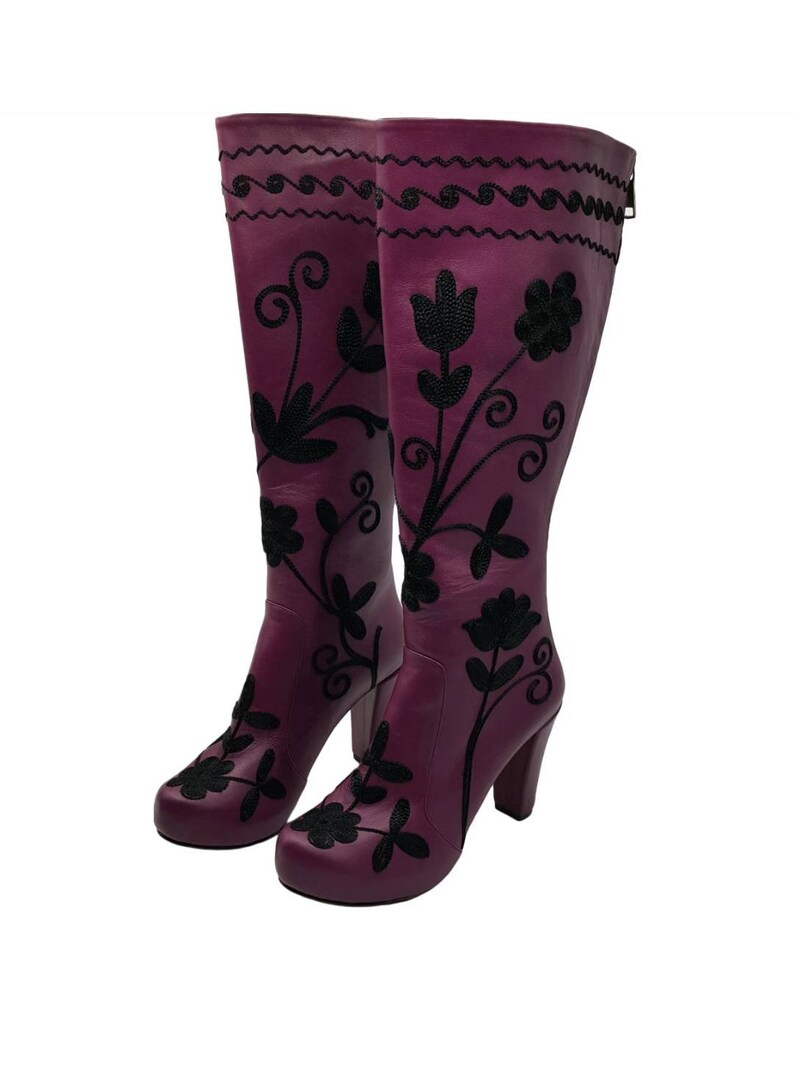 Puede incluir: Un par de botas altas de cuero morado con bordados florales negros y dise&ntilde;os de enredaderas. Las botas tienen punta redondeada, tac&oacute;n de bloque y un cierre de cremallera vertical en la parte posterior de la ca&ntilde;a.