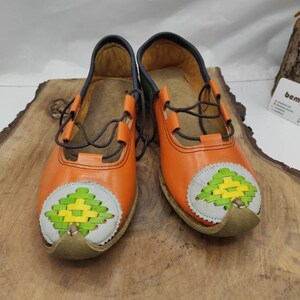 K&ouml;nnte beinhalten: Ein Paar orangefarbene Lederschuhe mit gr&uuml;nen und gelben gewebten Mustern an den Zehen. Die Schuhe haben braune Schn&uuml;rsenkel und ein dunkelblaues Futter.