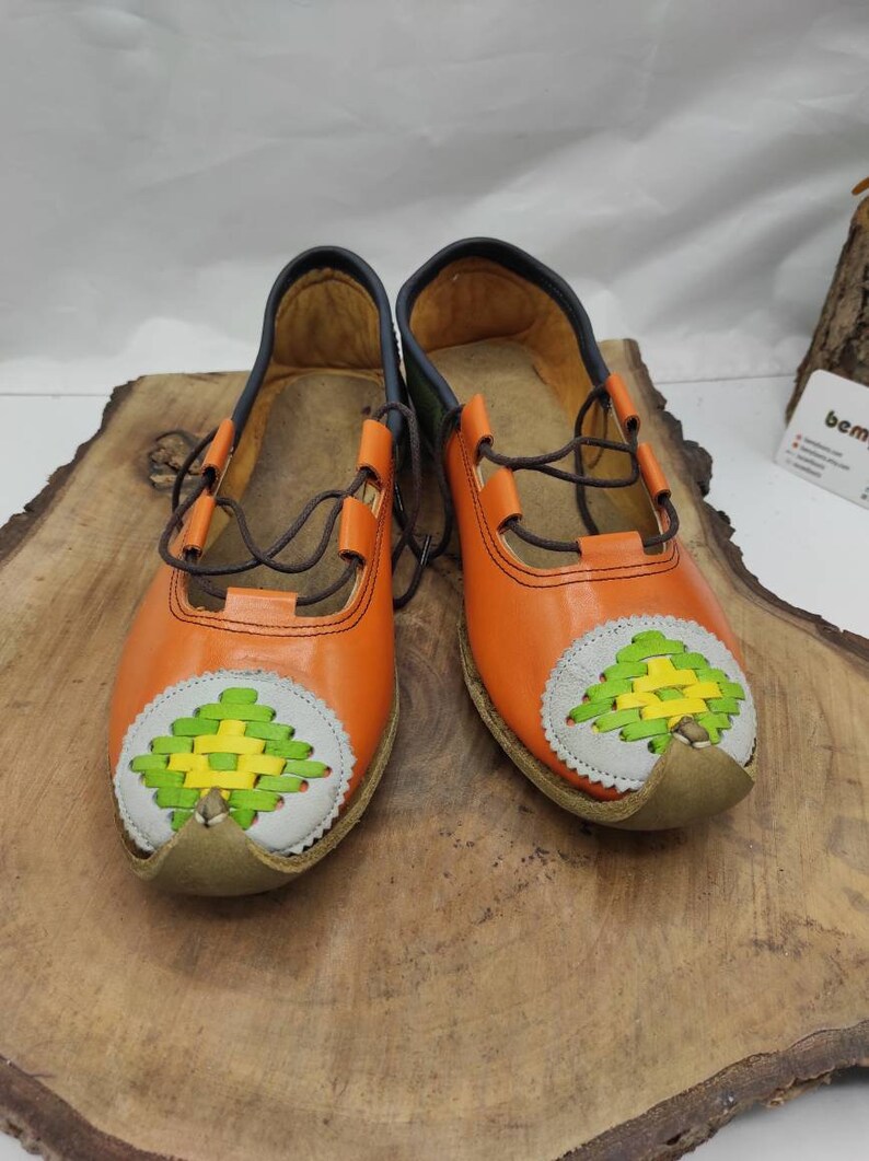 K&ouml;nnte beinhalten: Ein Paar orangefarbene Lederschuhe mit gr&uuml;nen und gelben gewebten Mustern an den Zehen. Die Schuhe haben braune Schn&uuml;rsenkel und ein dunkelblaues Futter.