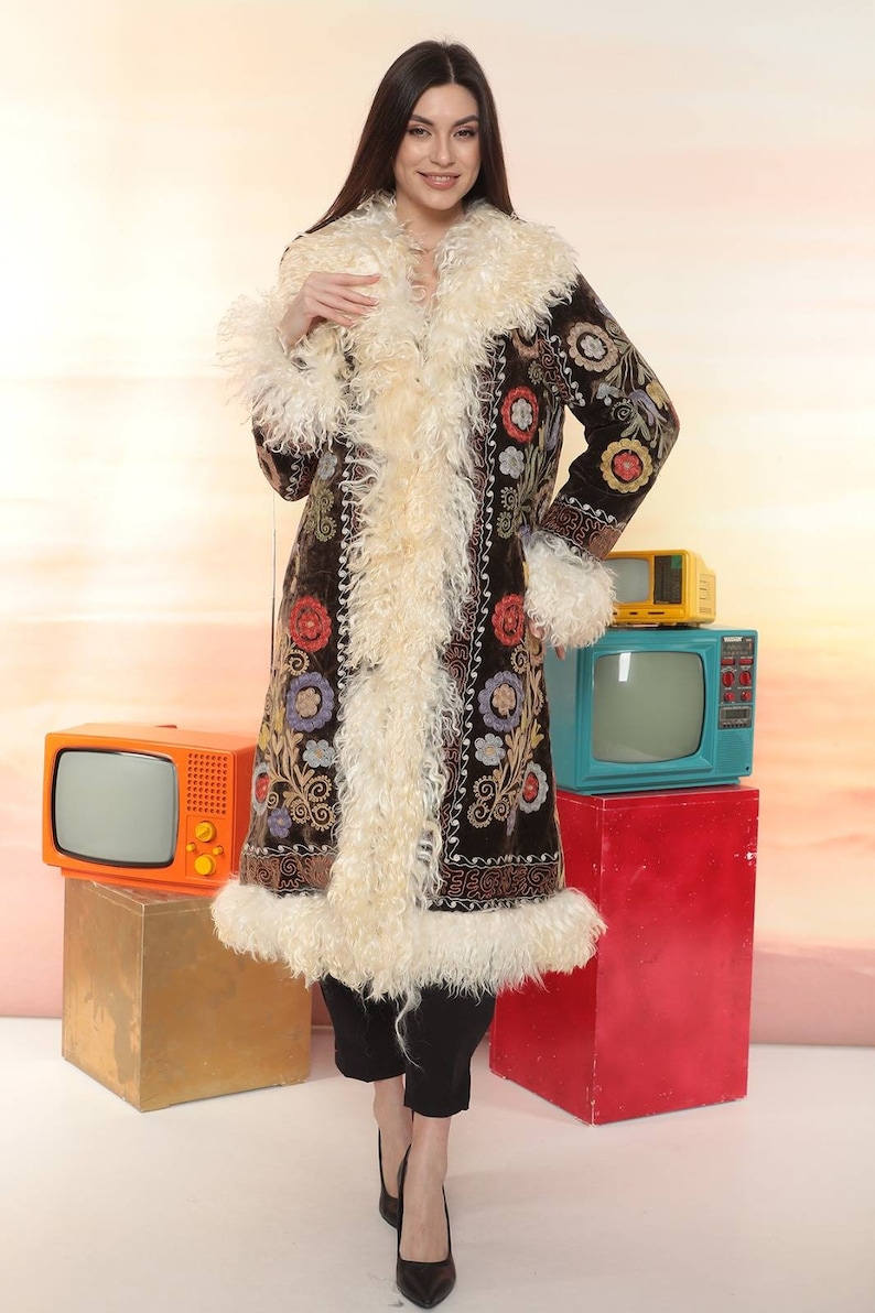 May include: A long, dark brown coat with colorful floral embroidery and cream-colored fur trim. The coat is worn over black pants and heels. Vintage televisions in orange, yellow, and teal are in the background.