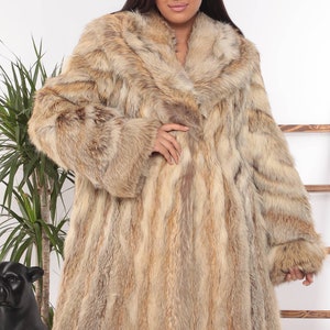 May include: A long, luxurious fur coat in shades of tan, brown, and cream. The coat features a wide collar and long sleeves. The model is wearing black pants. The coat appears to be made of genuine fur.