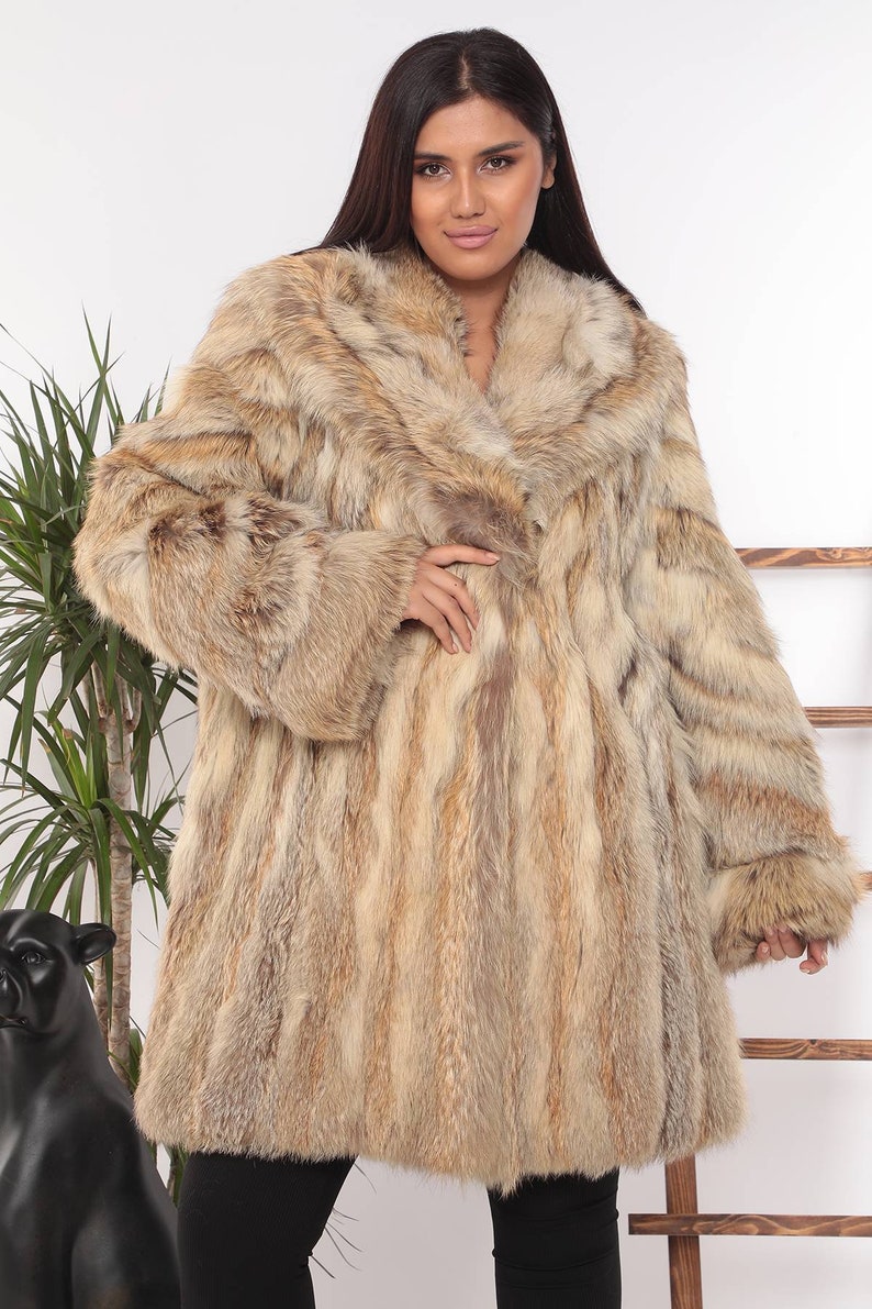 May include: A long, luxurious fur coat in shades of tan, brown, and cream. The coat features a wide collar and long sleeves. The model is wearing black pants. The coat appears to be made of genuine fur.
