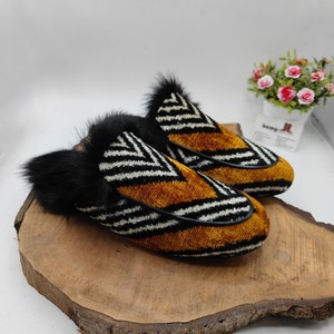 Puede incluir: Un par de zapatillas rayadas en blanco y negro con terciopelo marr&oacute;n y ribete de piel negra. Las zapatillas est&aacute;n sobre una superficie de madera.