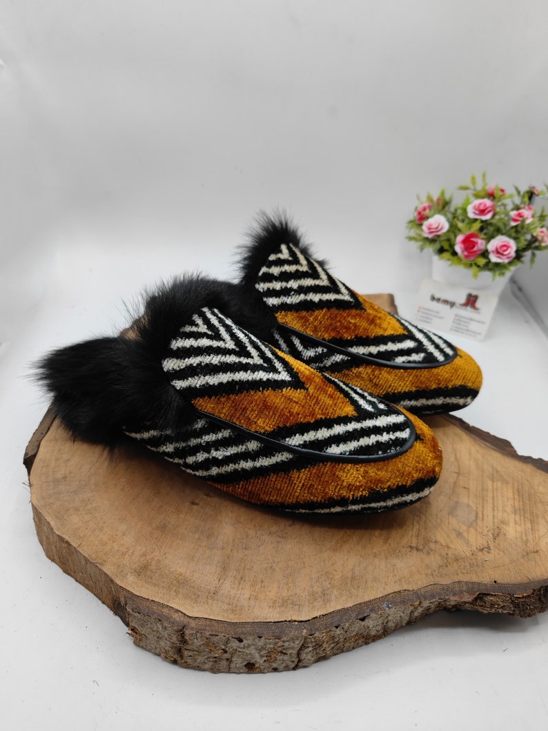 Puede incluir: Un par de zapatillas rayadas en blanco y negro con terciopelo marr&oacute;n y ribete de piel negra. Las zapatillas est&aacute;n sobre una superficie de madera.