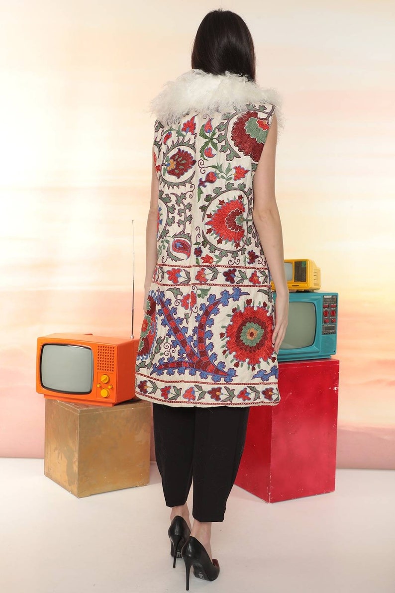 May include: A white sleeveless vest with a colorful floral embroidery pattern. The vest has a cream fur collar. The vest is worn over black pants.