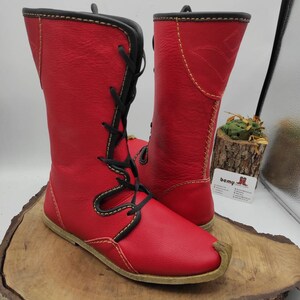 May include: A pair of red leather boots with black laces and yellow stitching. The boots have a pointed toe and a flat sole.