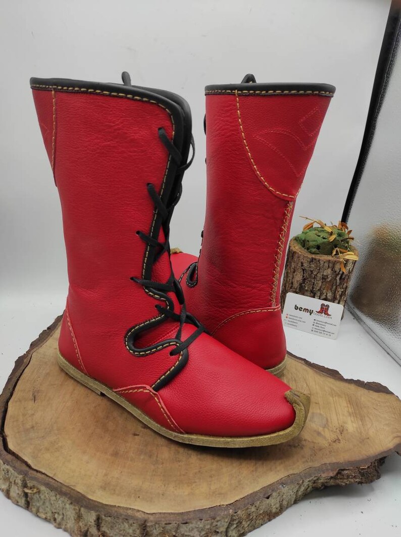 May include: A pair of red leather boots with black laces and yellow stitching. The boots have a pointed toe and a flat sole.