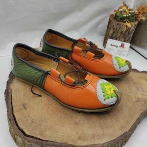 K&ouml;nnte beinhalten: Ein Paar orangefarbene und gr&uuml;ne Lederschuhe mit einem wei&szlig;en und gr&uuml;nen Stickmuster an der Spitze. Die Schuhe haben einen braunen Schn&uuml;rverschluss.