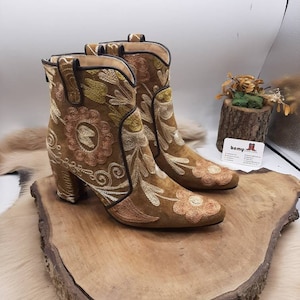May include: Brown suede ankle boots with floral embroidery in shades of pink, yellow, and green. The boots have a block heel and a pointed toe.