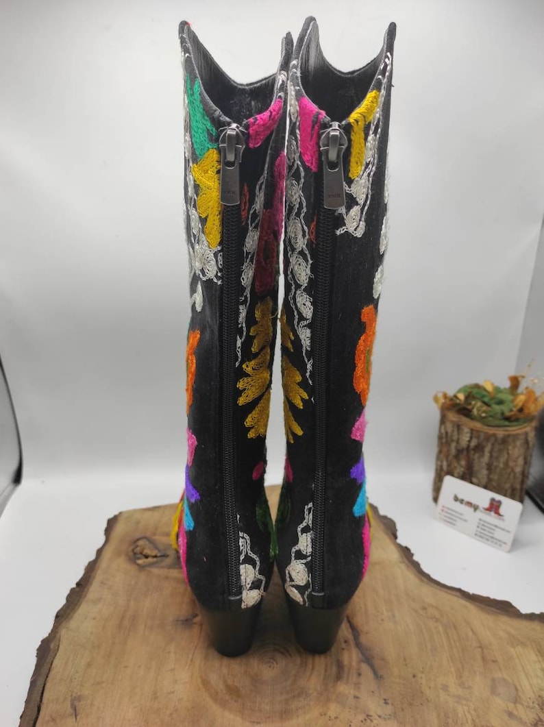 May include: A pair of black suede boots with colorful embroidery. The boots have a zipper on the inside of each boot and a block heel.