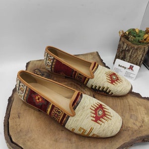 May include: A pair of flat, slip-on shoes with a woven textile design. The shoes feature a beige base with red, brown, and yellow geometric patterns. They have a leather trim and are displayed on a wooden surface.