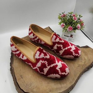 May include: A pair of red and white patterned velvet loafers with a brown leather trim. The loafers are sitting on a wooden surface.