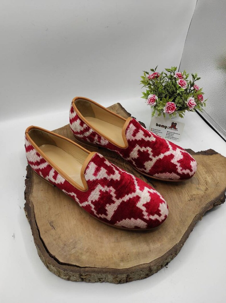 May include: A pair of red and white patterned velvet loafers with a brown leather trim. The loafers are sitting on a wooden surface.