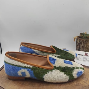 May include: A pair of blue, green, and white patterned velvet slippers with a brown leather trim. The slippers have a low heel and are sitting on a wooden surface.