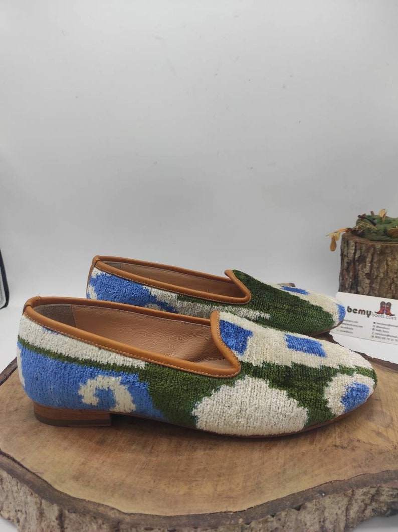 May include: A pair of blue, green, and white patterned velvet slippers with a brown leather trim. The slippers have a low heel and are sitting on a wooden surface.