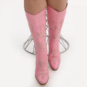 May include: A pair of pink leather cowboy boots with floral embroidery. The boots have a pointed toe and a high heel.