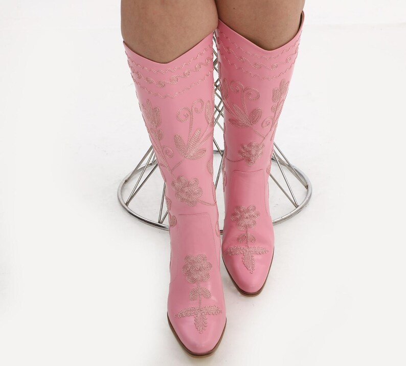 May include: A pair of pink leather cowboy boots with floral embroidery. The boots have a pointed toe and a high heel.