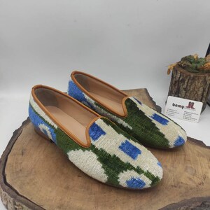 May include: A pair of white, green, and blue patterned velvet slippers with brown leather trim. The slippers are on a wooden surface.