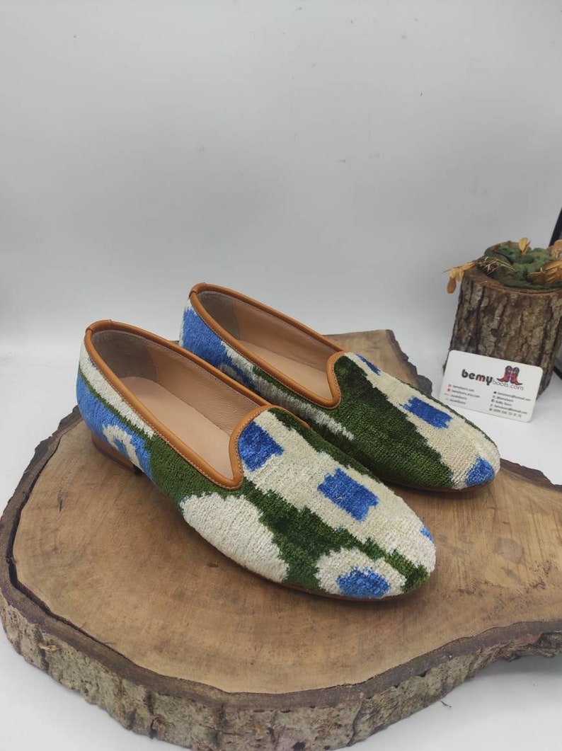 May include: A pair of white, green, and blue patterned velvet slippers with brown leather trim. The slippers are on a wooden surface.