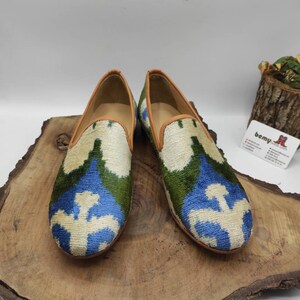 May include: A pair of blue, green, and white patterned velvet loafers with brown leather trim. The loafers are on a wooden surface.