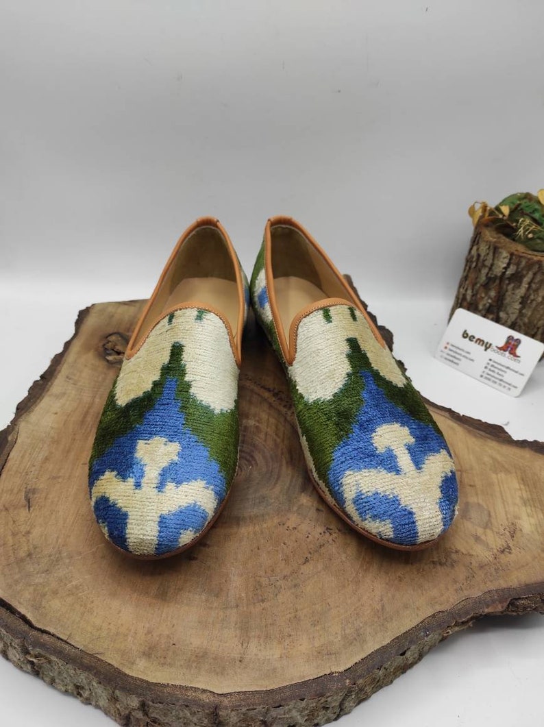 May include: A pair of blue, green, and white patterned velvet loafers with brown leather trim. The loafers are on a wooden surface.